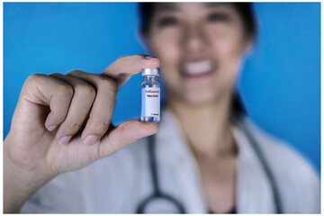A female doctor wearing a white coat with a stethoscope around her neck, holding a glass vial of Influenza vaccine or Flu shot with her fingers, smiling. Healthcare And Medical concept.