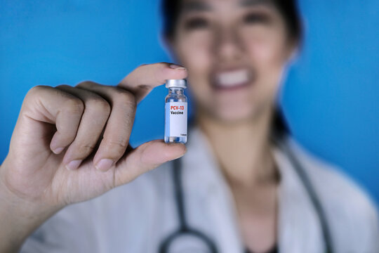 Female doctor wearing a white coat with a stethoscope around her shoulder holding a glass vial of PCV or Pneumococcal conjugate vaccine, smiling. Blue background. Healthcare And Medical concept.