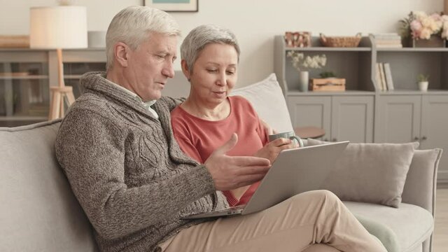 Medium long of cheerful senior Caucasian man hugging his Asian wife who holding mug, couple using laptop computer and talking, sitting on sofa at home