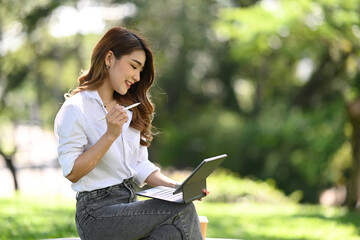 Pretty young woman sitting on bench in park and using digital tablet.