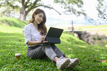 Happy young asian woman sitting on green grass in park and working with computer tablet.
