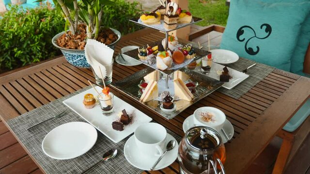 Traditional English afternoon tea stand with selection of sweets - cakes, snacks, sandwiches and macaroons. Tea set and coffee with assortment of pastries in outdoor restaurant. Sweet buffet breakfast