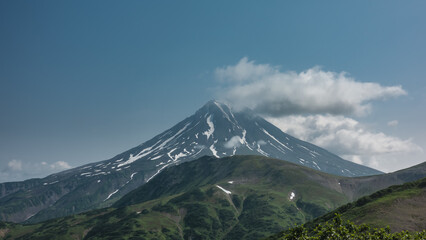 A beautiful conical volcano on a blue sky background. Snow on the slopes. Clouds at the top. In the foreground are green hills. Kamchatka. Stratovolcano Vilyuchinsky.