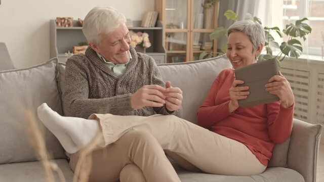 Medium long of short-haired Asian woman using tablet computer, leaning on couch with legs up on lap of senior Caucasian husband who fooling around with her and tickling her feet