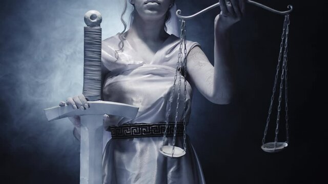 Goddess of justice holds sword and raises the scales. White woman on black background with smoke in studio. Closeup. Concept of legal justice