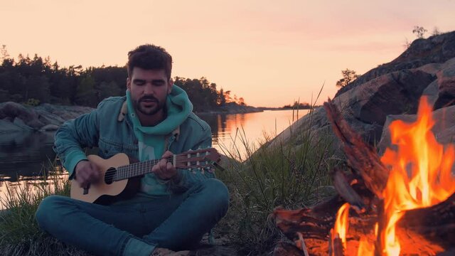 Young good looking Musician playing guitar and singing in front of bonfire on island at the ocean. Musician singing in front of friends and enjoy sunset while traveling. Filmed in ProRes.