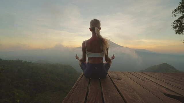 Fit woman in sportswear meditating on platform with view of Mount Agung, sunset