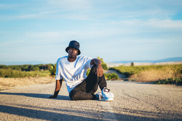 Young black handsome man wearing a bucket hat sitting on the ground in the middle of a road, sunset © Fran Martínez/Wirestock