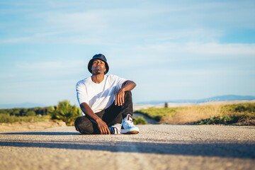 Young black handsome man wearing a bucket hat sitting on the ground in the middle of a road, sunset