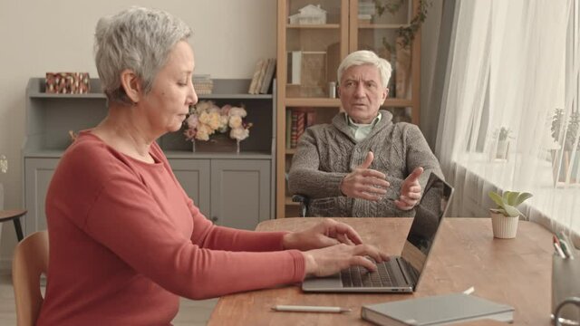 Side View Of Short-haired Asian Woman Sitting At Desk At Home, Typing On Portable Computer, Older Caucasian Man Dictating To Her