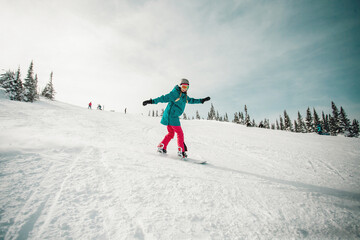 Young woman wearing blue and pink clothes snowboarding at almost empty tracks of Sheregesh ski resort, on a beautiful clear winter day. Female snowboarder in a motion.