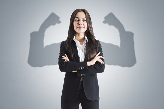 Portrait of attractive young european businesswoman with folded arms and shadow muscle arms on concrete wall background. Strenght and leadership concept.