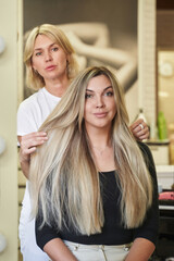 the hairdresser straightens in front of the mirror very long beautiful blond hair of a woman who smiles