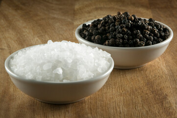 Black pepper corns and salt crystals in small white bowls on timber table