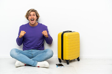 Young blonde man with suitcase sitting on the floor celebrating a victory in winner position