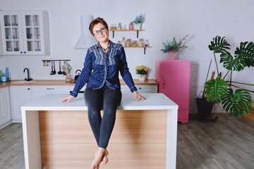 Young cute woman 30+ with short hair in blue jeans and a shirt sitting and relaxing in her bright clean kitchen. 