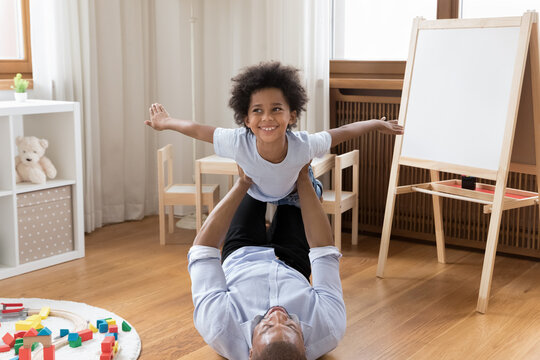 Happy young strong african american father lifting in air adorable little child son, lying on warm heated wooden floor in modern playroom, having fun playing airplane, imagining travelling at home.