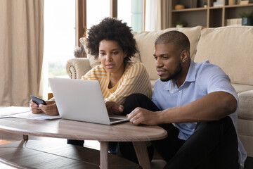 Concentrated young african american married couple looking at computer screen, analyzing household budget, paying bills in e-banking app, planning investment or managing budget together at home.