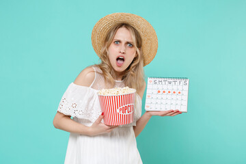 Shocked irritated young woman in white dress hat hold periods calendar for checking menstruation days bucket of popcorn isolated on blue turquoise background. Medical healthcare gynecological concept.