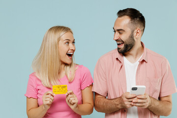 Young smiling couple two friends family man woman in casual clothes use mobile cell phone credit bank card shopping online booking tour together isolated on pastel plain light blue background studio.