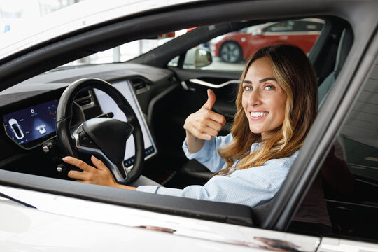 Woman customer buyer client in blue shirt hold put hands on steering wheel show thumb up choose auto want buy new automobile in car showroom vehicle salon dealership motor show indoor Sales concept