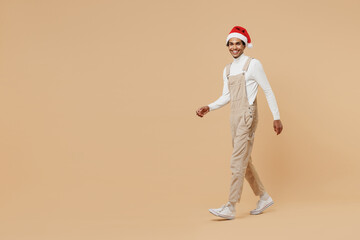 Fototapeta na wymiar Full body smiling fun happy young african man 20s wear Santa Claus red Christmas hat walking going isolated on plain pastel beige background studio portrait. Happy New Year 2022 celebration concept.