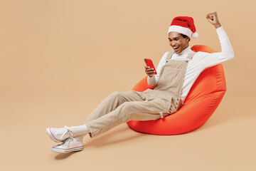 Full size young african man in red Christmas hat sit in bag chair hold mobile cell phone do winner...