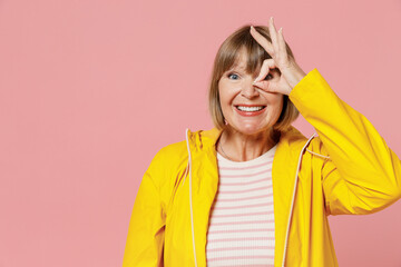Mature elderly woman 55 years old wears yellow waterproof raincoat show hide eye with okay ok gesture isolated on plain pastel light pink background studio portrait. Outdoors wet fall weather concept.