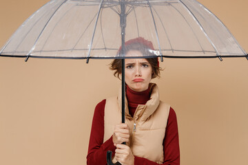 Young upset frowning unhappy woman in red turtleneck vest beret hold transparent umbrella look camera isolated on plain pastel beige background studio portrait. People lifestyle rainy weather concept. - obrazy, fototapety, plakaty