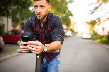 Young urban man driving his scooter in the park holding his phone typing a text, wearing checked shirt and a watch, enjoying in the park, looking up to the sky