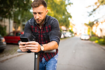 Young urban man driving his scooter in the park holding his phone typing a text, wearing checked shirt and a watch, enjoying in the park