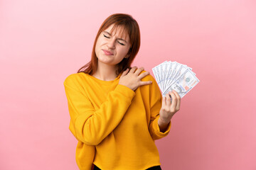 Redhead girl taking a lot of money isolated on pink background suffering from pain in shoulder for having made an effort