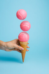 Woman's hand holding ice cream cone with pink tennis balls on pastel blue background. Creative...