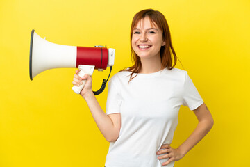 Redhead girl isolated on yellow background holding a megaphone and smiling