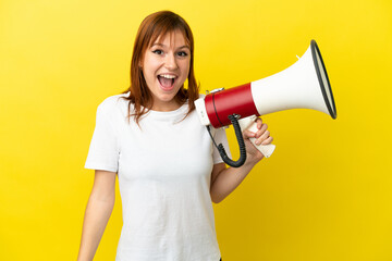 Redhead girl isolated on yellow background holding a megaphone and with surprise expression