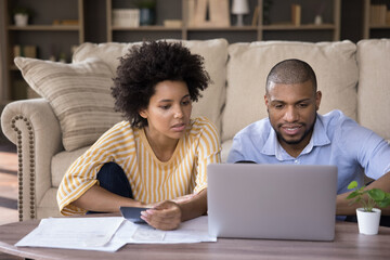 Focused young african american family couple calculating expenses, analyzing paper bills, paying for services in e-banking computer application, managing monthly budget or planning investment at home.
