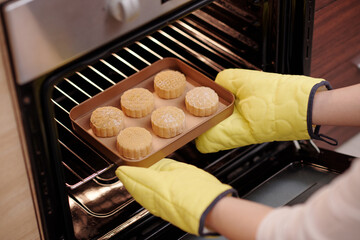 Woman in baking gloves putting tray with mooncakes in preheated oven when cooking delicious dessert...