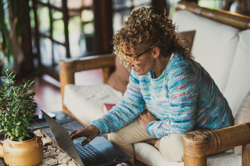 Adult woman with blue sweater  sitting on the couch at home working and using laptop computer to...