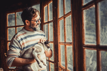 Adult happy mature man with a cat look outside the windows at home enjoying indoor leisure relax...