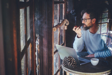 Mature hipster man use computer laptop at home in chalet wooden house smoking pipe and tobacco...