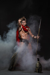 A young athletic man dressed as a Roman soldier with a red cloak stands with a spear in his hand in full growth on a black background shrouded in mysterious smoke. - obrazy, fototapety, plakaty
