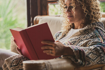 Relaxed adult lady reading a book sitting on the chair at home with outside view. Concept of female people happy lifestyle living alone. Attracvite woman studying indoor - Powered by Adobe