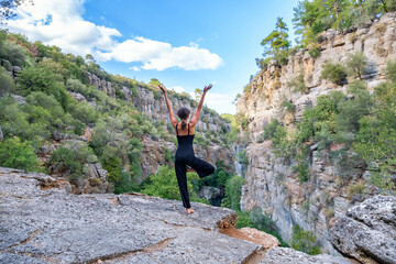 Full body back view of anonymous female practicing Vrikshasana posture while standing on one leg with raised arms on edge of rocky cliff