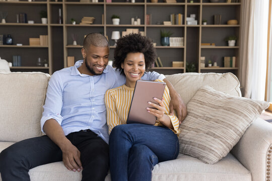 Happy young married african american family couple using digital computer tablet, choosing goods shopping in internet store, web surfing information, spending time online, sitting on sofa at home.