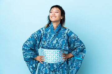 Young woman wearing kimono over isolated blue background posing with arms at hip and smiling