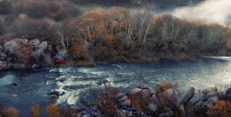 Panoramic photo of the autumn landscape. A river with rapids and rocky banks and autumn trees on the bank in a foggy haze. Selective focus. Southern Bug River.