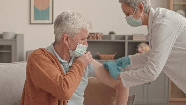 Medium of female Asian doctor wearing scrubs, gloves and mask giving injection to senior Caucasian man at daytime at home
