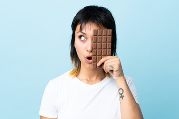 Young Uruguayan woman over isolated blue background taking a chocolate tablet and surprised
