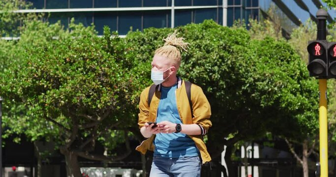 Albino african american man with face mask and dreadlocks walking and using smartphone