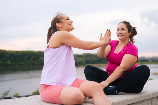 Cheerful slim fitness female trainer and motivated fat young woman greeting each other with a high-five other after exercising outdoors. Instructor help overweight woman lose weight outside.
